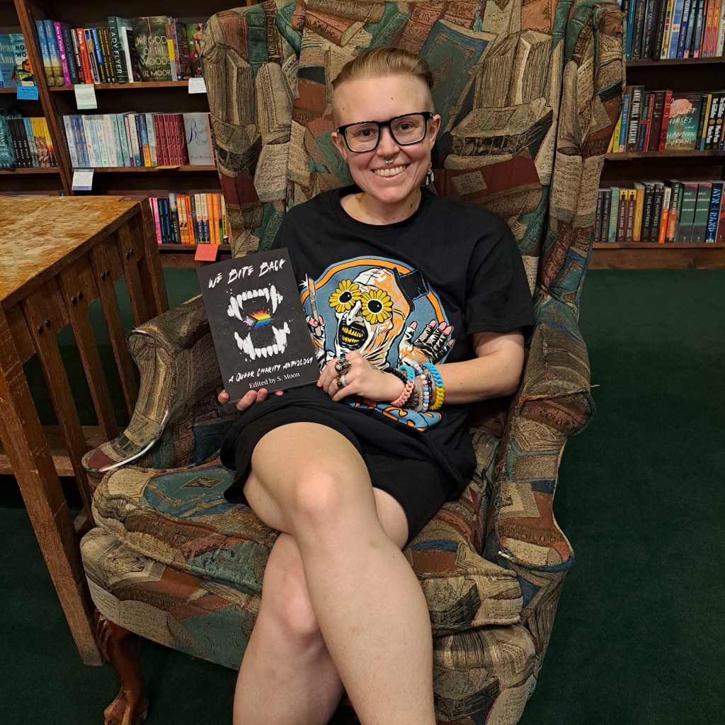 Sage Moon sitting on a chair holding "We Bite Back" at Tattered Cover Aspen Grove

